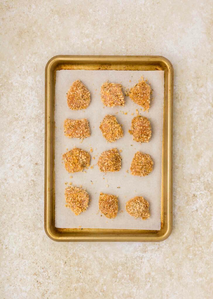 A baking tray lined with parchment paper holds twelve uncooked, breaded chicken nuggets. They are evenly spaced and placed on a light-colored countertop.