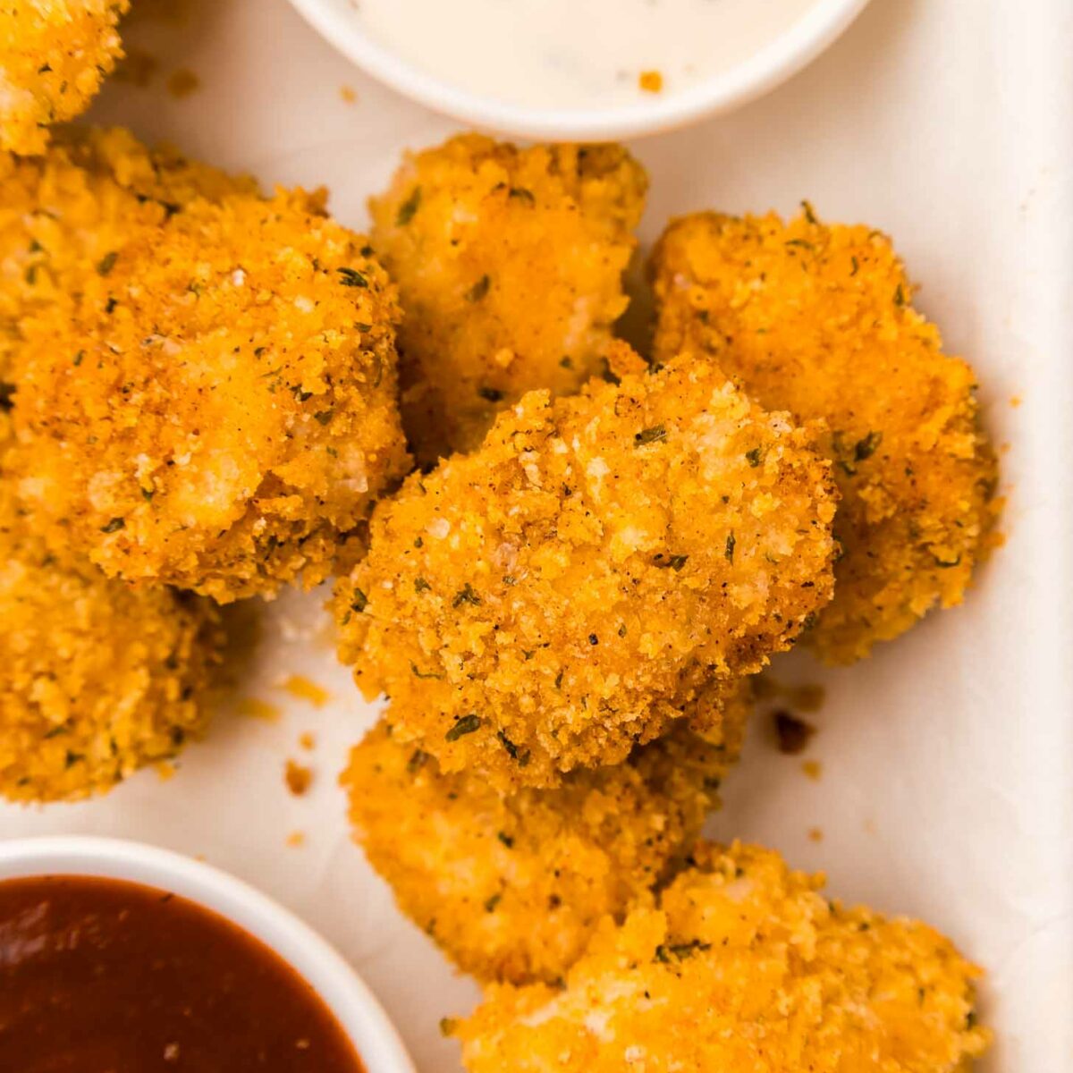 Golden, crispy chicken nuggets with a seasoned breadcrumb coating are arranged next to two dipping sauces; one is a creamy white ranch and the other is a rich, dark red bbq sauce. The scene is set on a white surface.