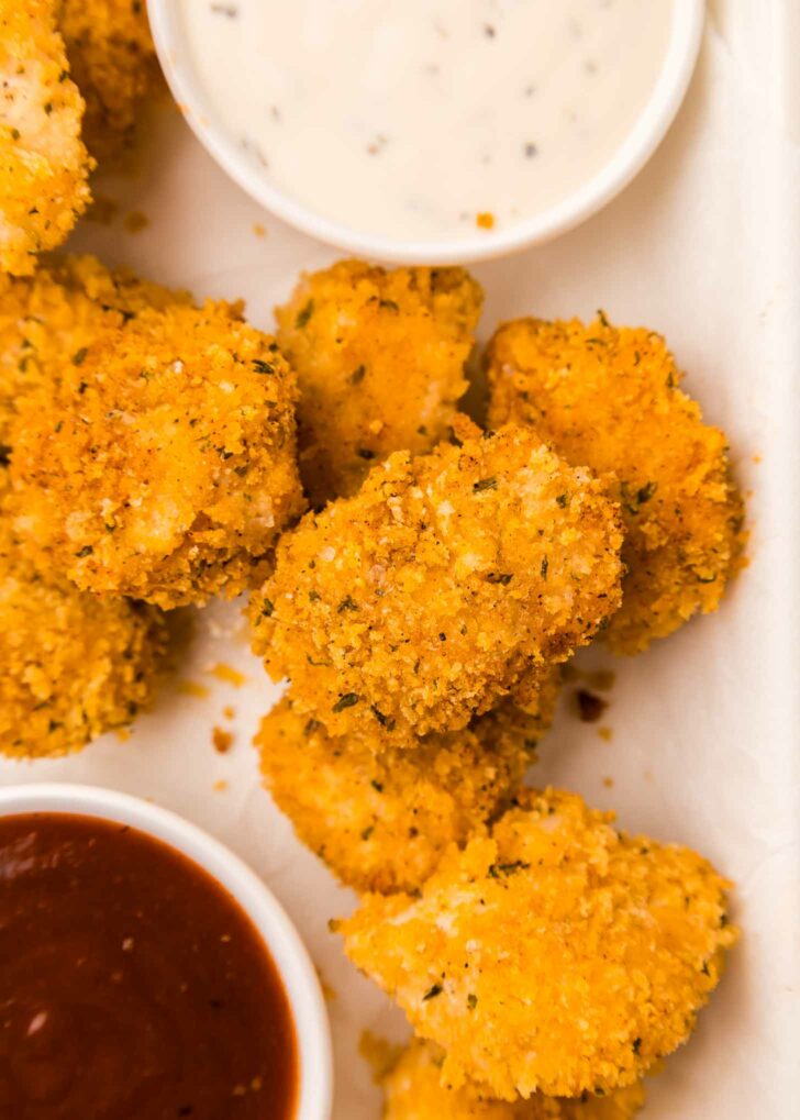 Golden, crispy chicken nuggets with a seasoned breadcrumb coating are arranged next to two dipping sauces; one is a creamy white ranch and the other is a rich, dark red bbq sauce. The scene is set on a white surface.
