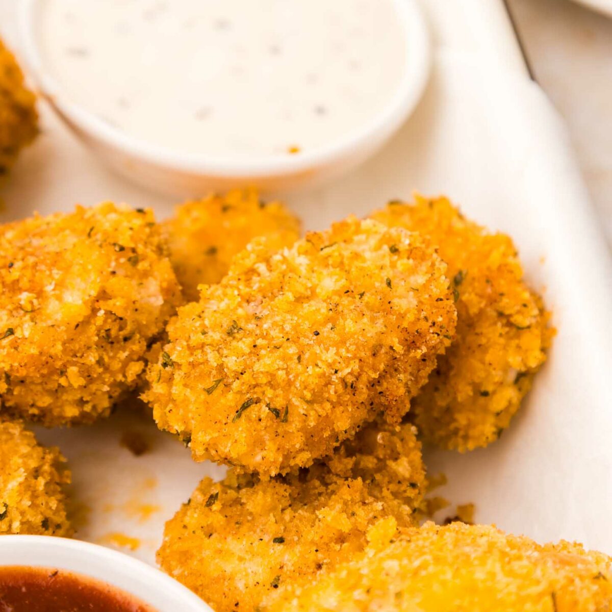 Close-up of crispy, golden-brown chicken nuggets on a white plate, alongside small bowls of ranch and barbecue dipping sauces. The texture of the nuggets is crunchy, with visible herbs in the breading.