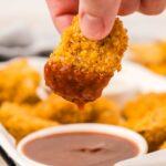 A hand dips a crispy chicken nugget into a bowl of barbecue sauce. In the background, more nuggets are visible on a white tray.
