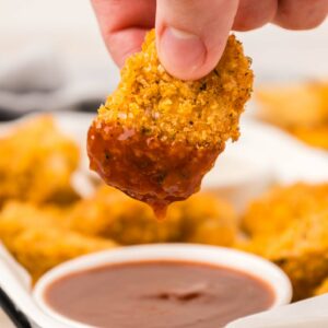 A hand dips a crispy chicken nugget into a bowl of barbecue sauce. In the background, more nuggets are visible on a white tray.