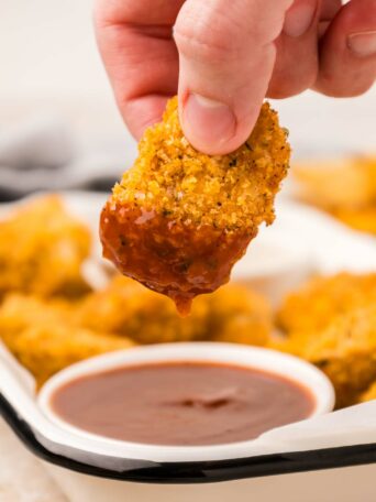 A hand dips a crispy chicken nugget into a bowl of barbecue sauce. In the background, more nuggets are visible on a white tray.