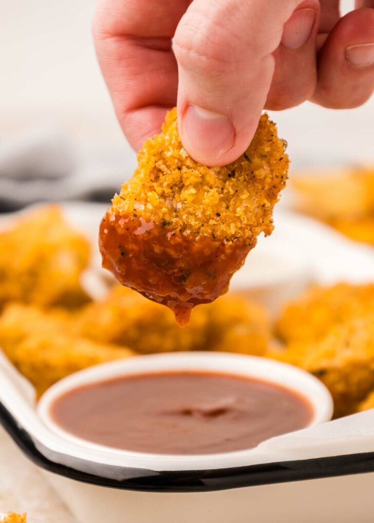 A hand dips a crispy chicken nugget into a bowl of barbecue sauce. In the background, more nuggets are visible on a white tray.