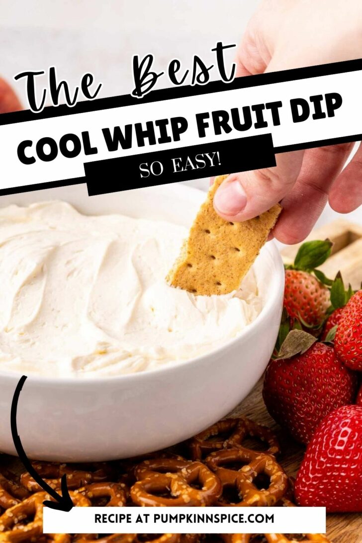 A hand dips a graham cracker into a bowl of creamy cool whip fruit dip. Strawberries and pretzels are arranged nearby. Text on the image reads: The Best Cool Whip Fruit Dip—So Easy! Recipe at pumpkinnspice.com.