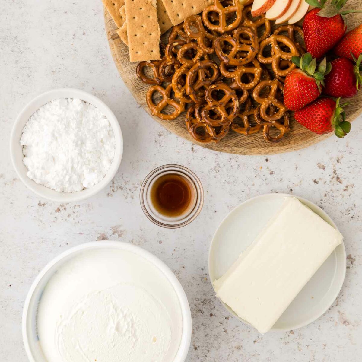 A wooden board with graham crackers, apple slices, pretzels, and strawberries next to bowls of powdered sugar, vanilla extract, cream cheese, and a bowl of cool whip on a light surface.