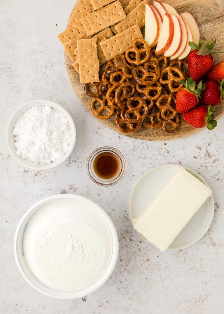 A wooden board with graham crackers, apple slices, pretzels, and strawberries next to bowls of powdered sugar, vanilla extract, cream cheese, and a bowl of cool whip on a light surface.