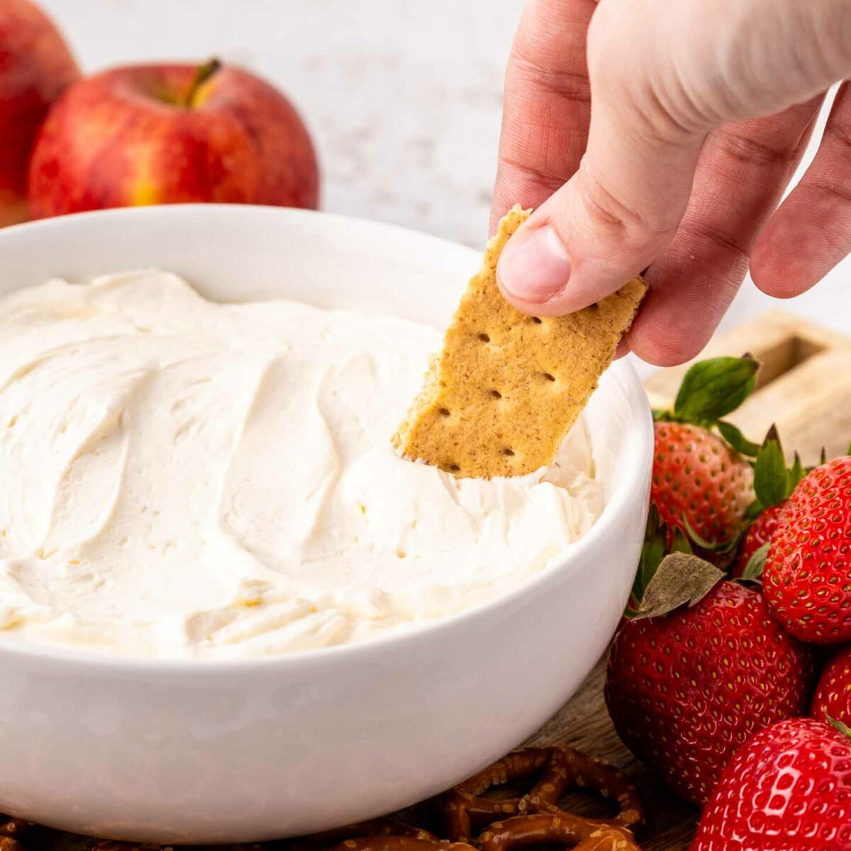 A hand dips a graham cracker into a bowl of creamy cool whip dip, surrounded by fresh strawberries, apples, and pretzels on a wooden board.