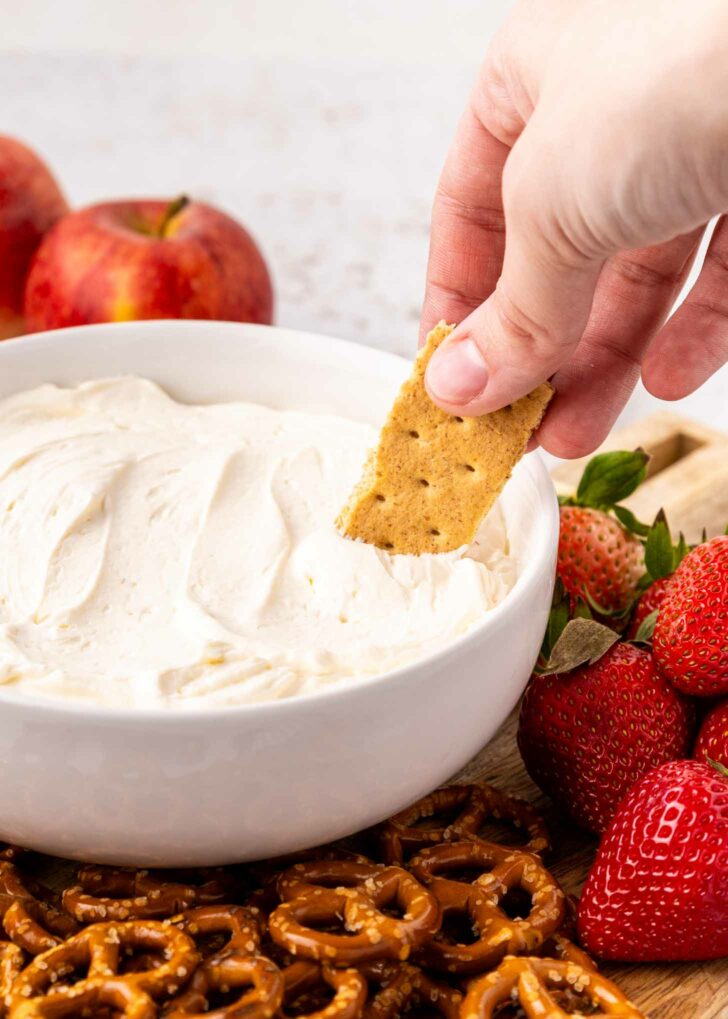 A hand dips a graham cracker into a bowl of creamy cool whip dip, surrounded by fresh strawberries, apples, and pretzels on a wooden board.