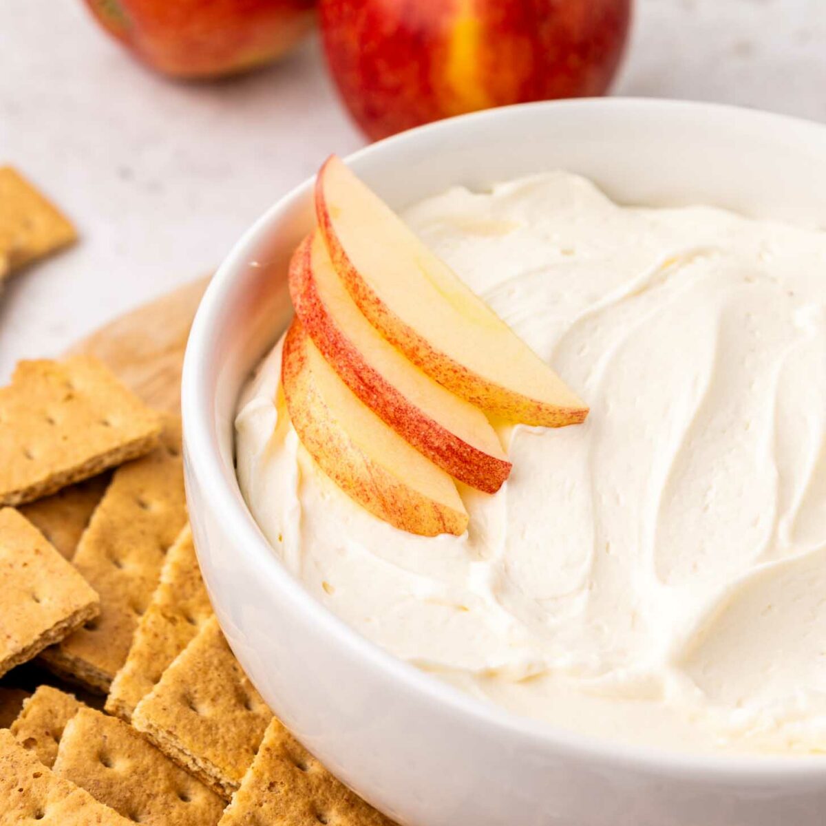 A white bowl filled with creamy cool whip dip, topped with apple slices, is surrounded by graham crackers, pretzels, and two whole apples in the background.