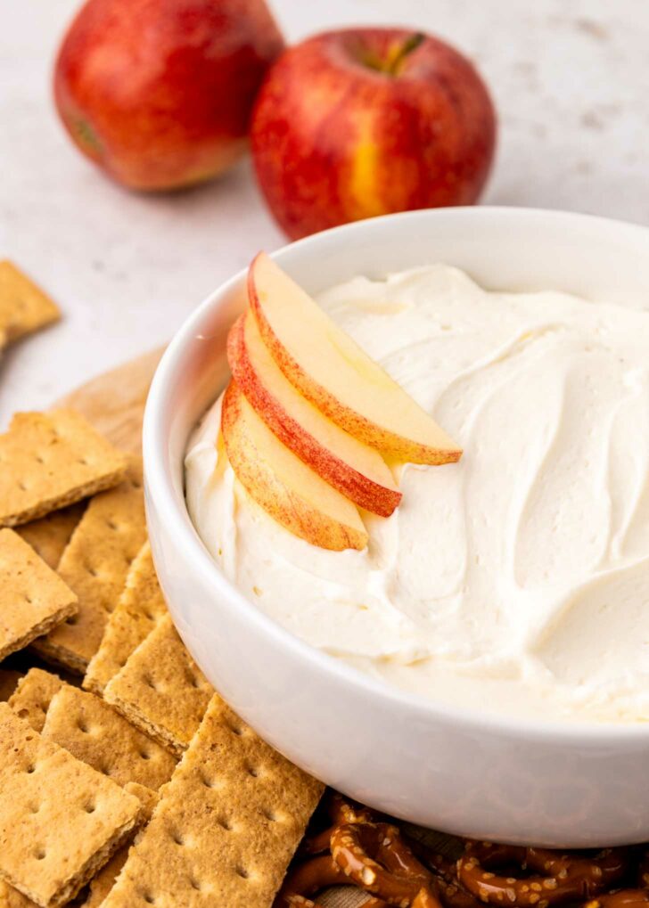 A white bowl filled with creamy cool whip dip, topped with apple slices, is surrounded by graham crackers, pretzels, and two whole apples in the background.
