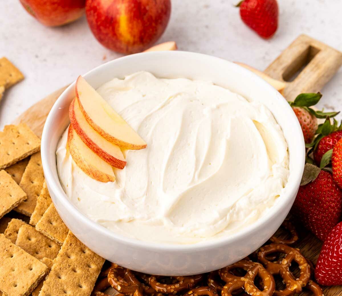A bowl of creamy cool whip dip topped with apple slices is surrounded by graham crackers, pretzels, fresh strawberries, and whole apples on a wooden board.