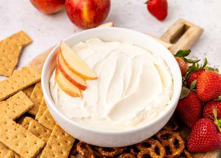 A bowl of creamy cool whip dip topped with apple slices is surrounded by graham crackers, pretzels, fresh strawberries, and whole apples on a wooden board.
