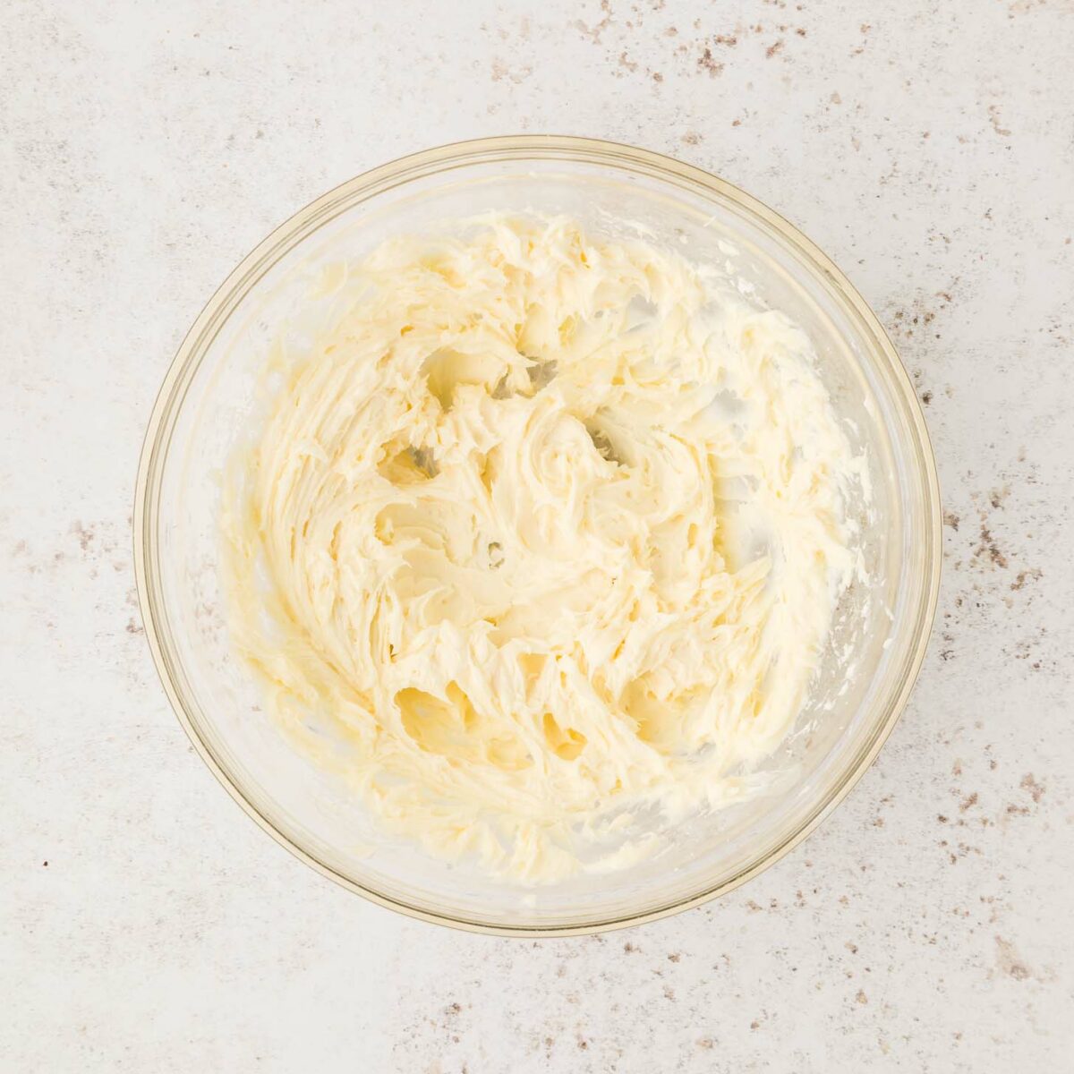 A glass bowl filled with creamy, cool whip, powdered sugar, vanilla, and cream cheese mixed together on a light, speckled countertop.