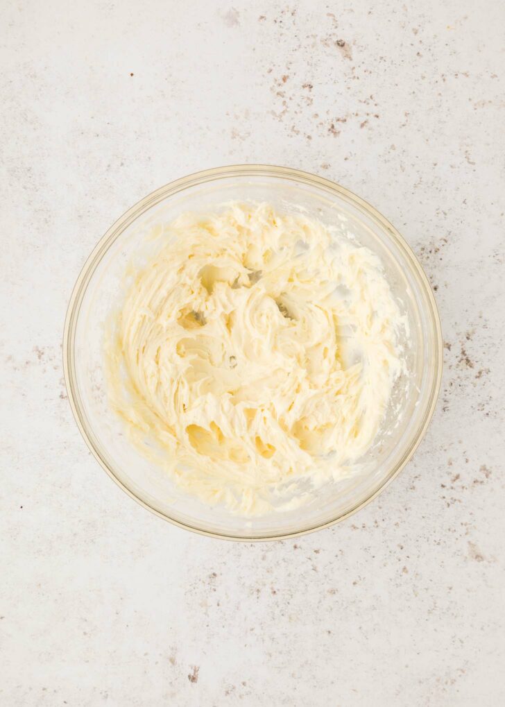 A glass bowl filled with creamy, cool whip, powdered sugar, vanilla, and cream cheese mixed together on a light, speckled countertop.