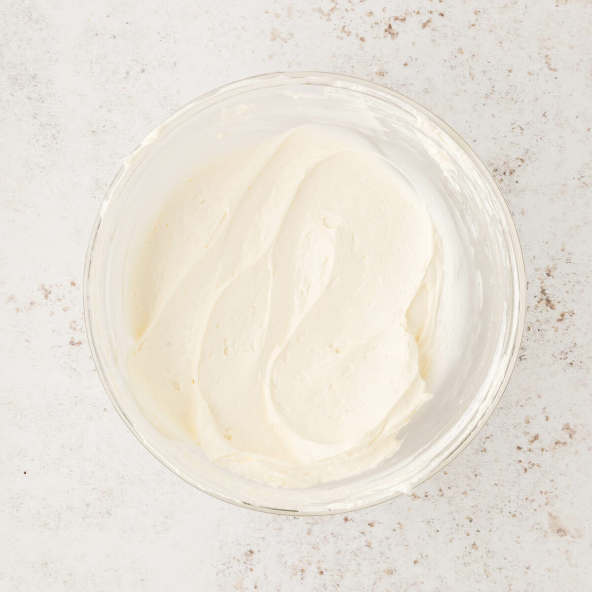 A glass bowl filled with smooth, creamy cool whip dip sits on a lightly speckled, off-white surface.