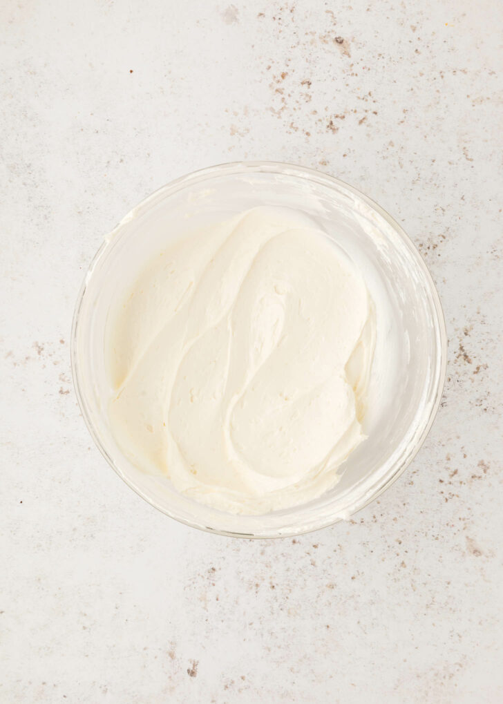 A glass bowl filled with smooth, creamy cool whip dip sits on a lightly speckled, off-white surface.