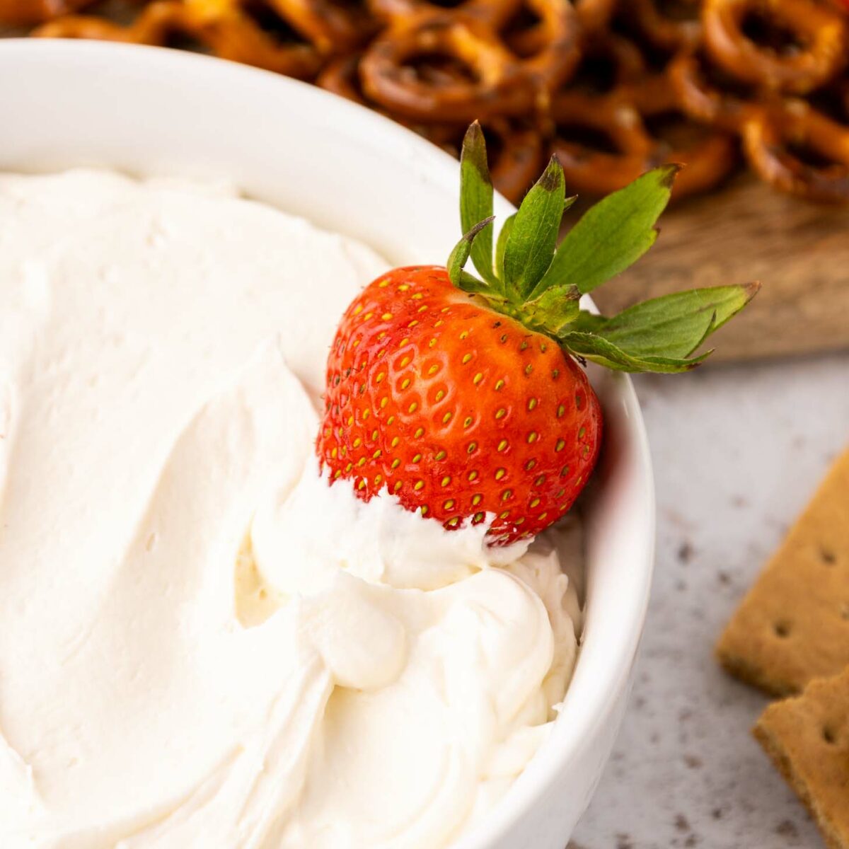 A fresh strawberry dipped in creamy white dip rests on the edge of a bowl, with pretzels and graham crackers arranged in the background.