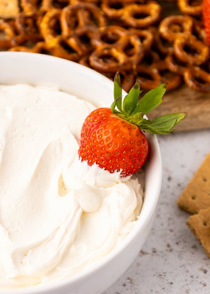 A fresh strawberry dipped in creamy white dip rests on the edge of a bowl, with pretzels and graham crackers arranged in the background.
