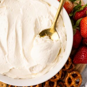 A bowl of creamy white dip with a gold spoon inside, surrounded by fresh strawberries, apple slices, pretzels, and graham crackers on a wooden board.