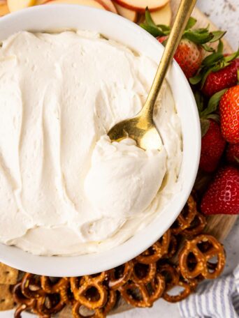 A bowl of creamy white dip with a gold spoon inside, surrounded by fresh strawberries, apple slices, pretzels, and graham crackers on a wooden board.