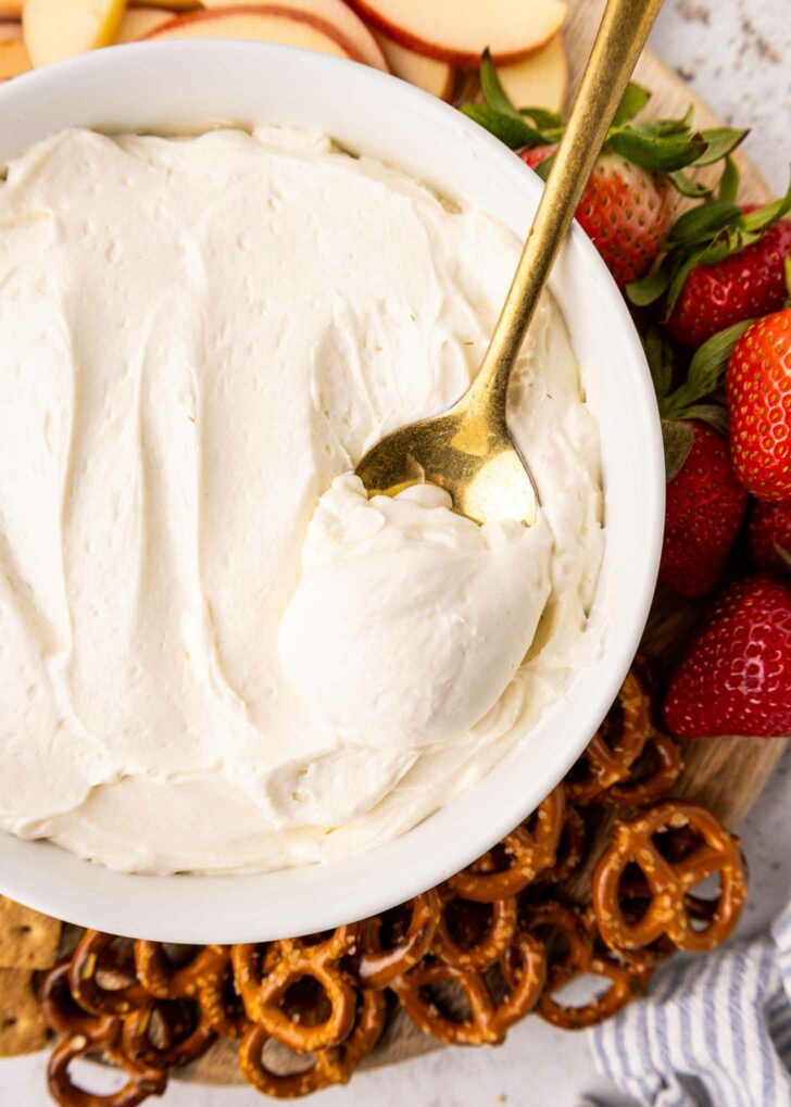 A bowl of creamy white dip with a gold spoon inside, surrounded by fresh strawberries, apple slices, pretzels, and graham crackers on a wooden board.