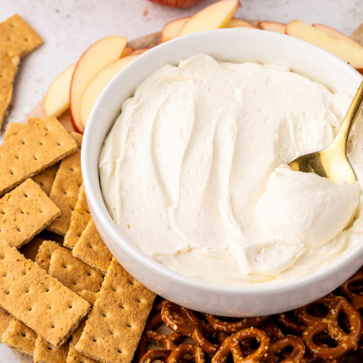 A bowl of creamy dip with a spoon is surrounded by sliced apples, graham crackers, and pretzels on a wooden board. Apples and a strawberry are in the background.