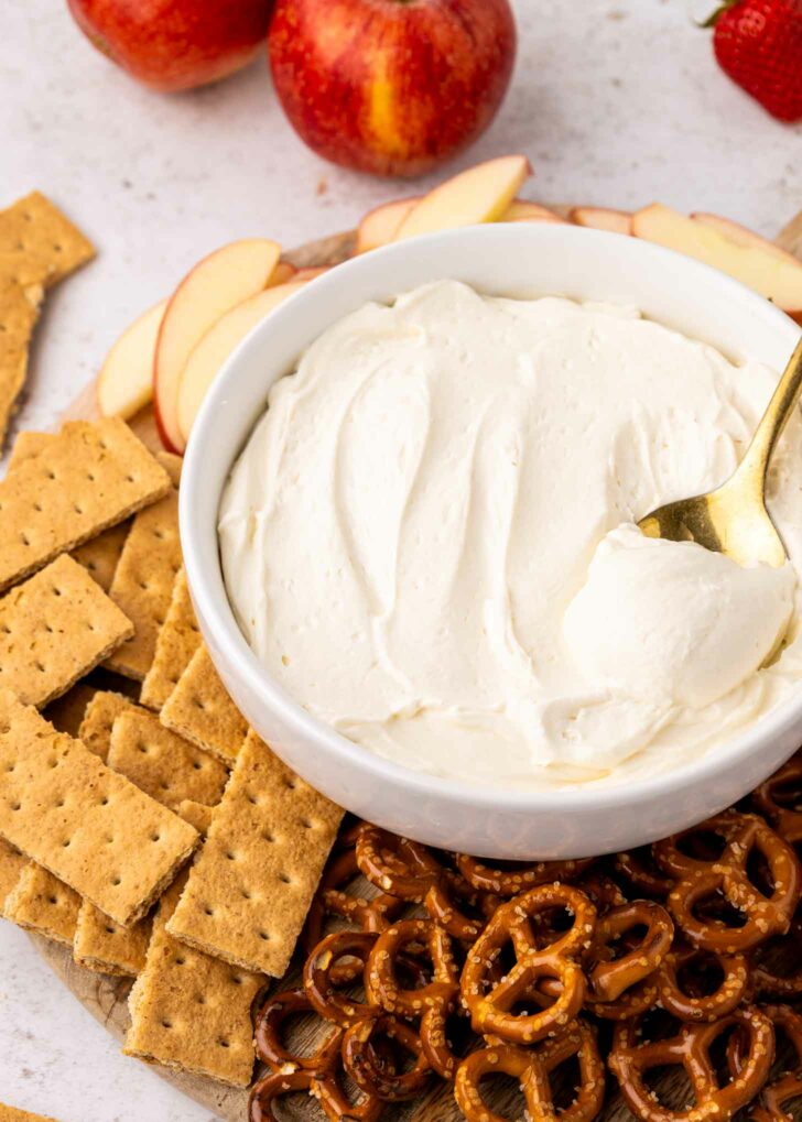A bowl of creamy dip with a spoon is surrounded by sliced apples, graham crackers, and pretzels on a wooden board. Apples and a strawberry are in the background.