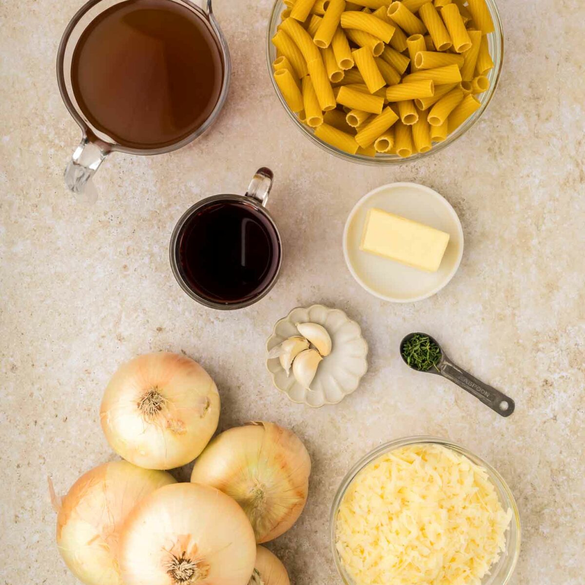 A top-down view of ingredients on a light counter: several yellow onions, dry pasta, a bowl of shredded cheese, a small bowl of butter, garlic cloves, a measuring spoon of thyme, red wine, and beef broth.