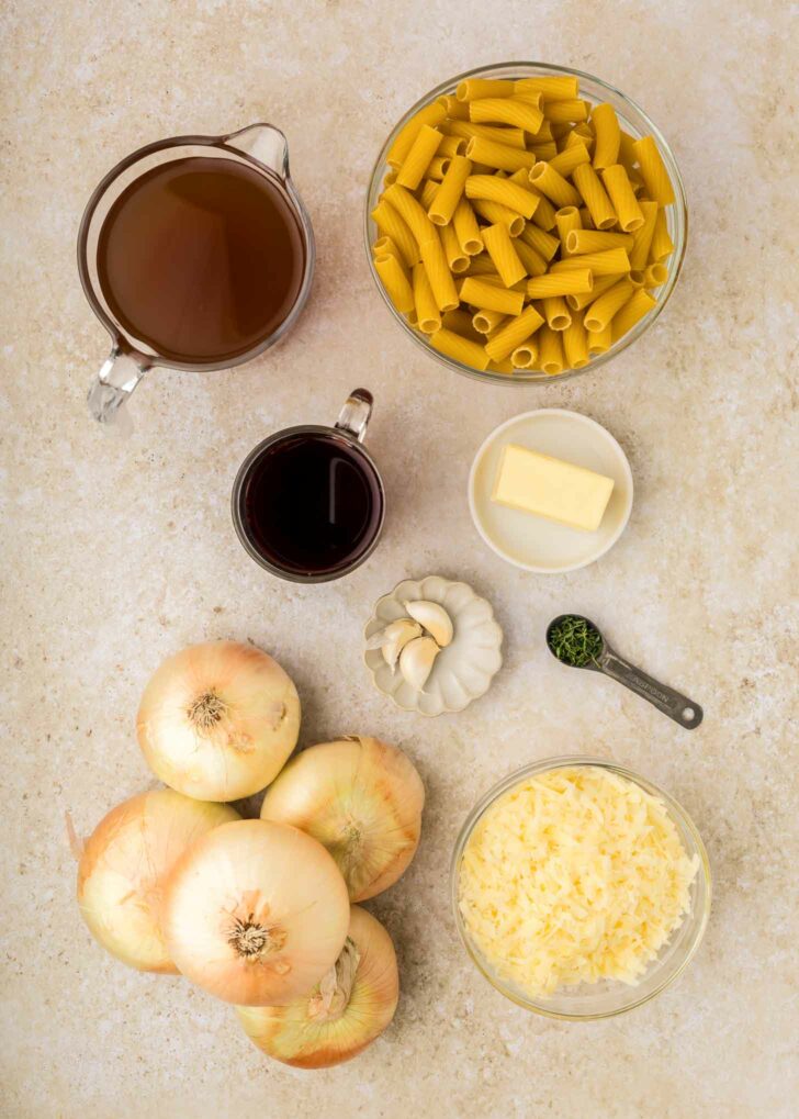 A top-down view of ingredients on a light counter: several yellow onions, dry pasta, a bowl of shredded cheese, a small bowl of butter, garlic cloves, a measuring spoon of thyme, red wine, and beef broth.