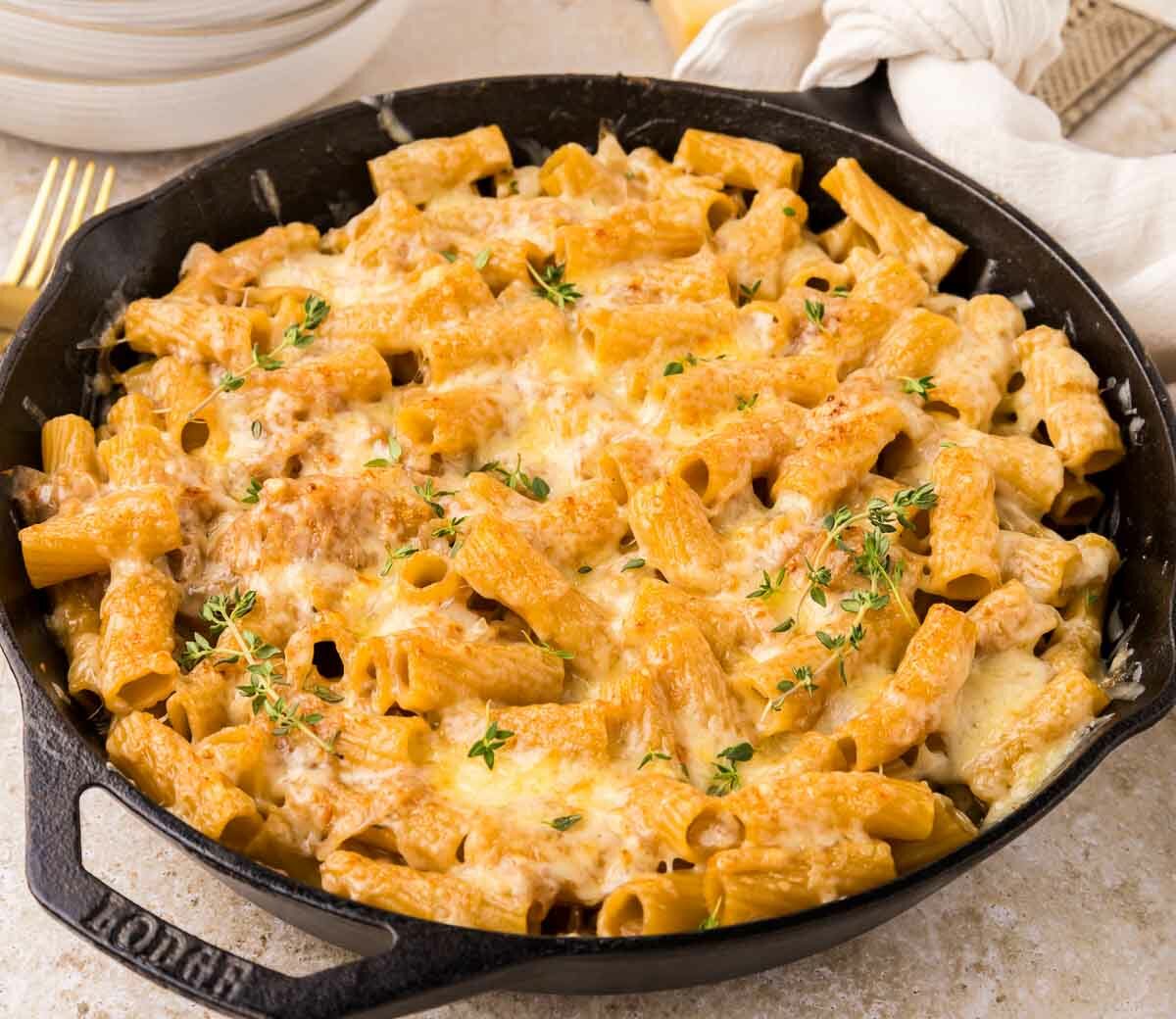 A cast iron skillet filled with baked rigatoni pasta covered in melted cheese and onions and garnished with fresh herbs, set on a light-colored table next to gold utensils and a white napkin.