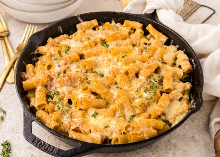 A cast iron skillet filled with baked rigatoni pasta covered in melted cheese and onions and garnished with fresh herbs, set on a light-colored table next to gold utensils and a white napkin.