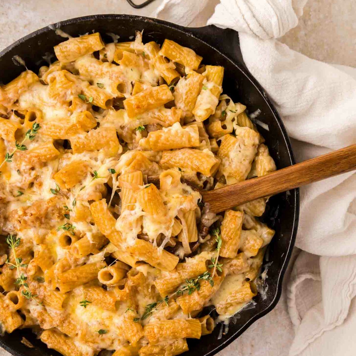 A skillet filled with rigatoni pasta topped with melted cheese and fresh thymne, with a wooden spoon resting inside and a white towel wrapped around the skillet handle.