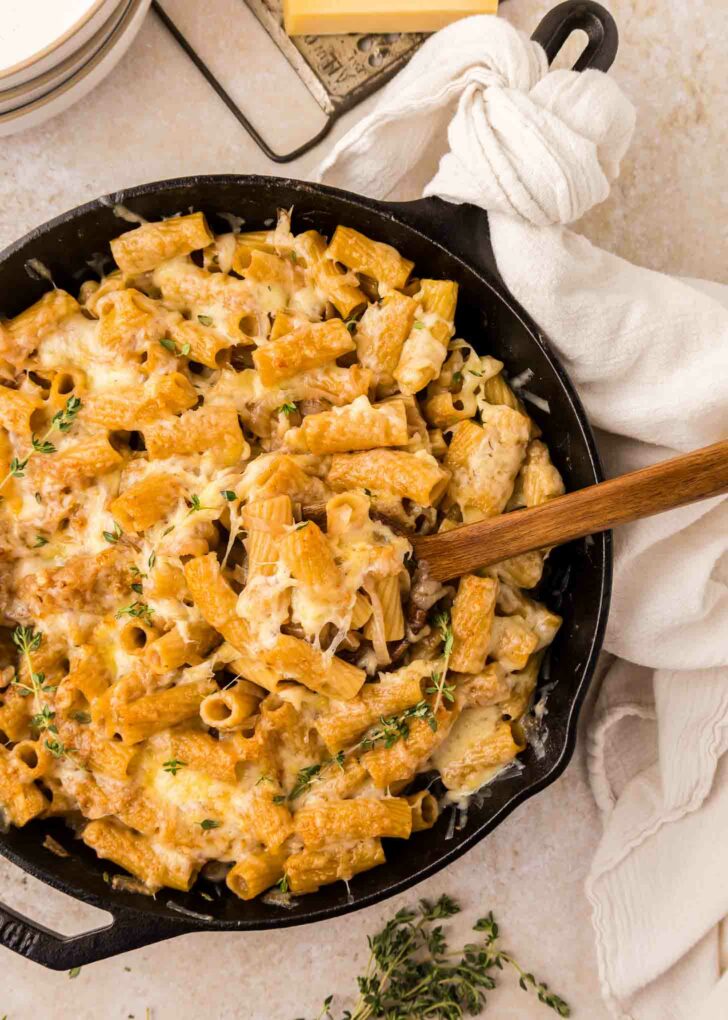 A skillet filled with rigatoni pasta topped with melted cheese and fresh thymne, with a wooden spoon resting inside and a white towel wrapped around the skillet handle.