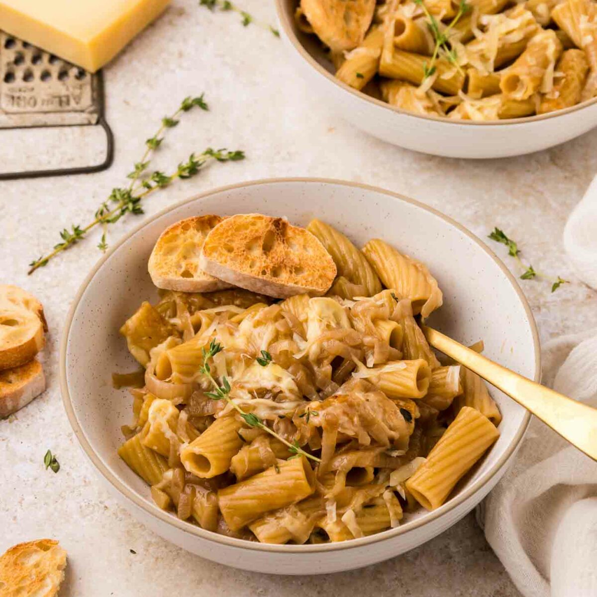 A bowl of rigatoni pasta topped with grated cheese, caramelized onions, fresh thyme, and toasted bread slices, with another bowl and cheese grater in the background on a light surface.