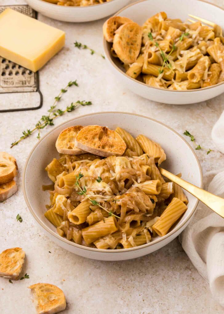A bowl of rigatoni pasta topped with grated cheese, caramelized onions, fresh thyme, and toasted bread slices, with another bowl and cheese grater in the background on a light surface.
