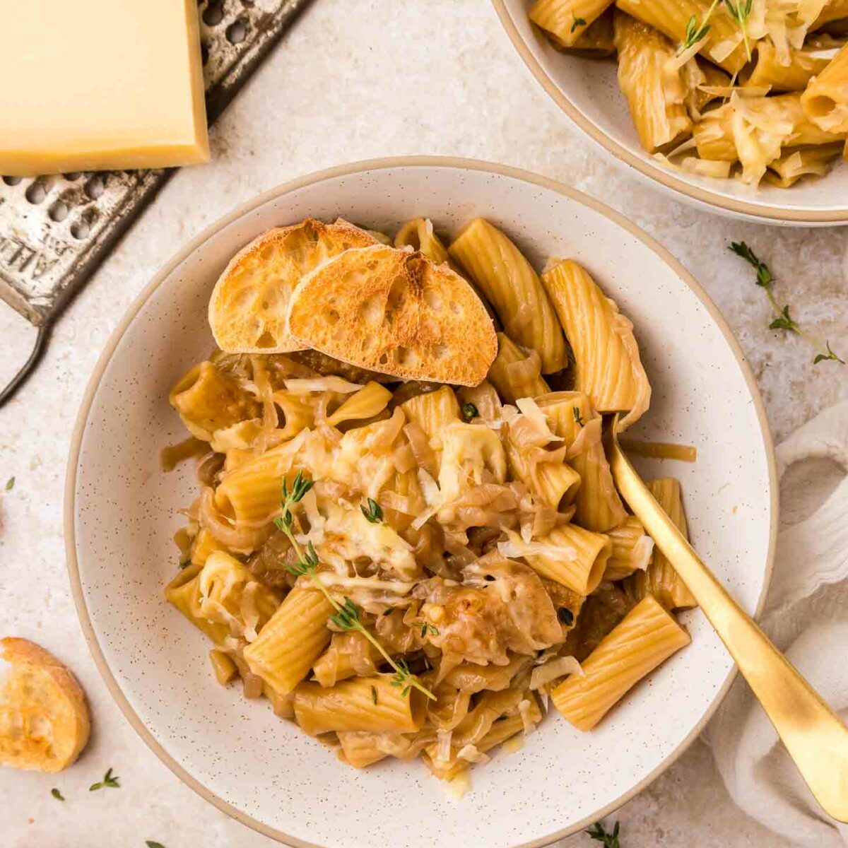 A bowl of rigatoni pasta topped with caramelized onions, grated cheese, fresh thyme, and slices of toasted baguette, with a gold fork beside it. Another bowl and cheese grater are visible in the background.