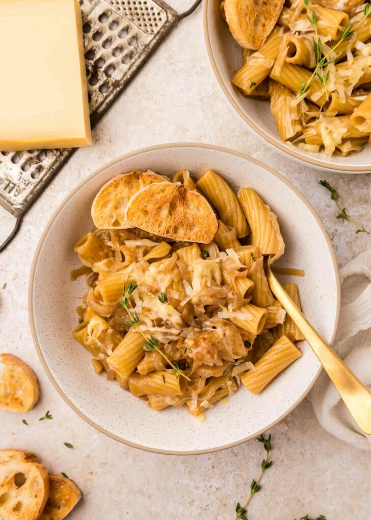 A bowl of rigatoni pasta topped with caramelized onions, grated cheese, fresh thyme, and slices of toasted baguette, with a gold fork beside it. Another bowl and cheese grater are visible in the background.