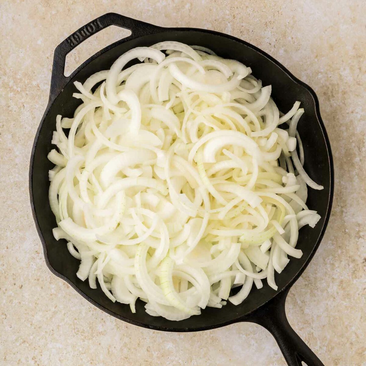 A cast iron skillet filled with raw, thinly sliced yellow onions sits on a light beige countertop.