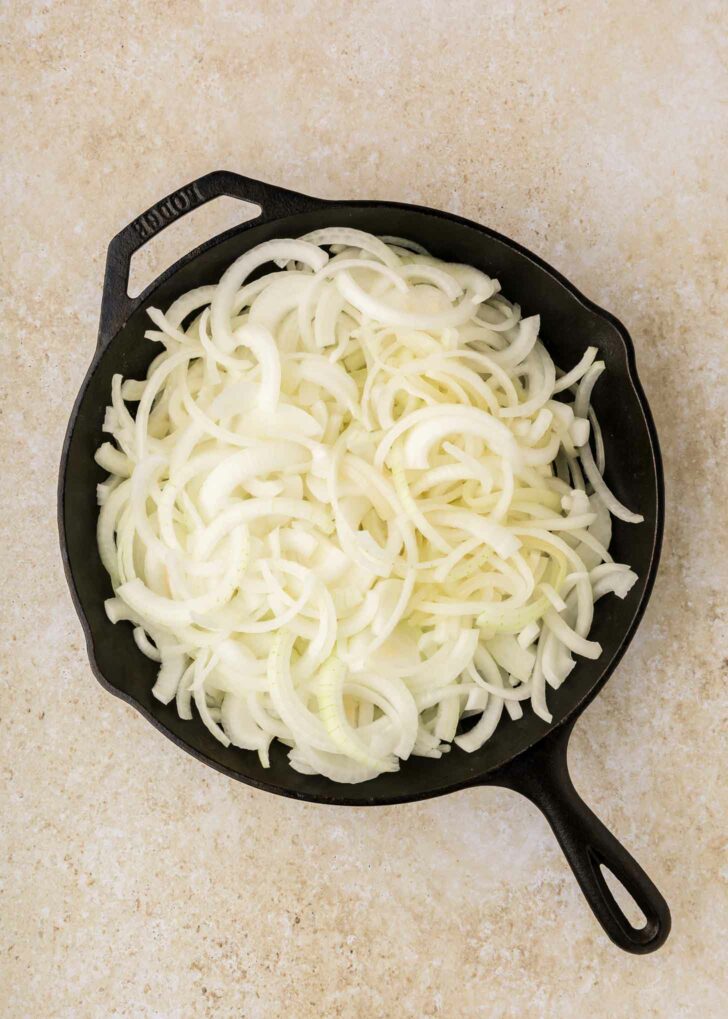 A cast iron skillet filled with raw, thinly sliced yellow onions sits on a light beige countertop.