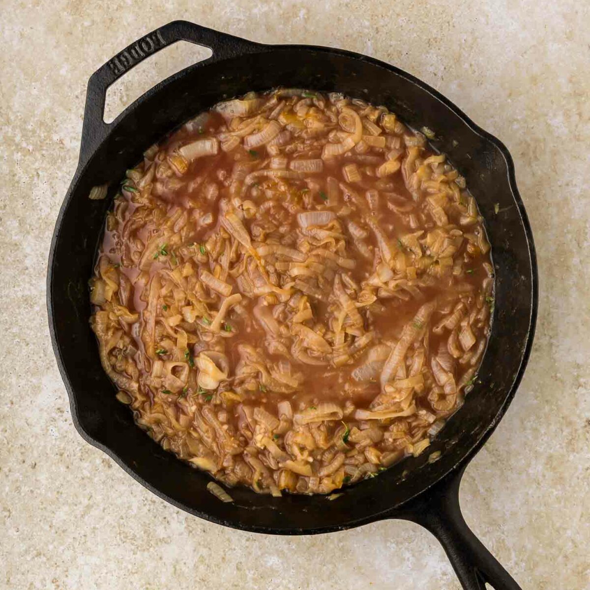 A cast iron skillet filled with caramelized onions simmering in a brown, brothy liquid, placed on a light beige countertop.