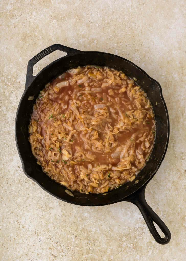 A cast iron skillet filled with caramelized onions simmering in a brown, brothy liquid, placed on a light beige countertop.
