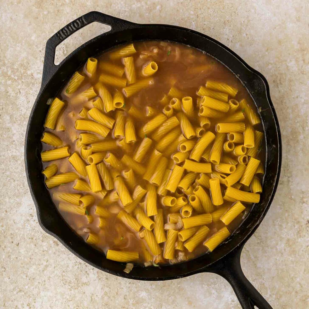 A black cast iron skillet filled with uncooked rigatoni pasta and brown liquid, set on a light beige countertop.