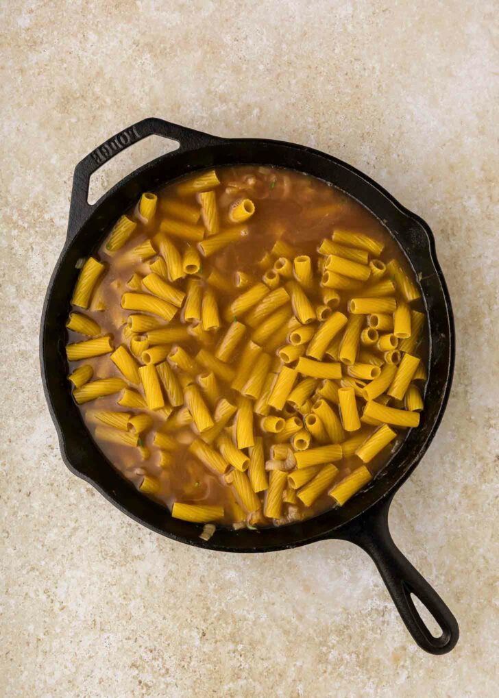 A black cast iron skillet filled with uncooked rigatoni pasta and brown liquid, set on a light beige countertop.