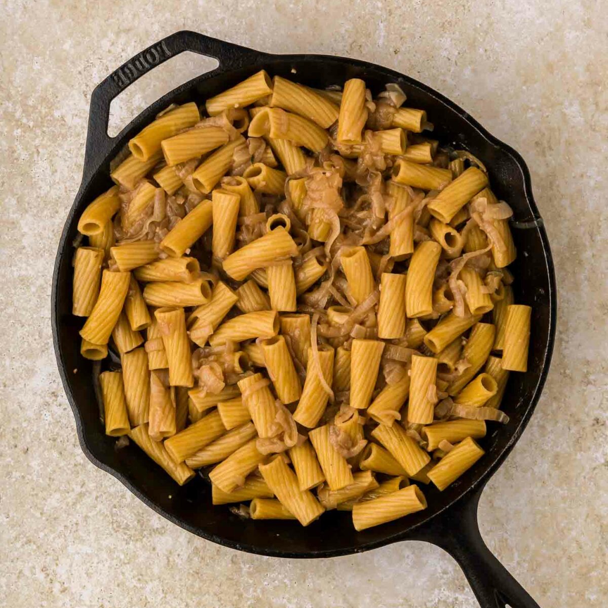 A black cast iron skillet filled with cooked rigatoni pasta mixed with caramelized onions, set on a light beige textured surface.