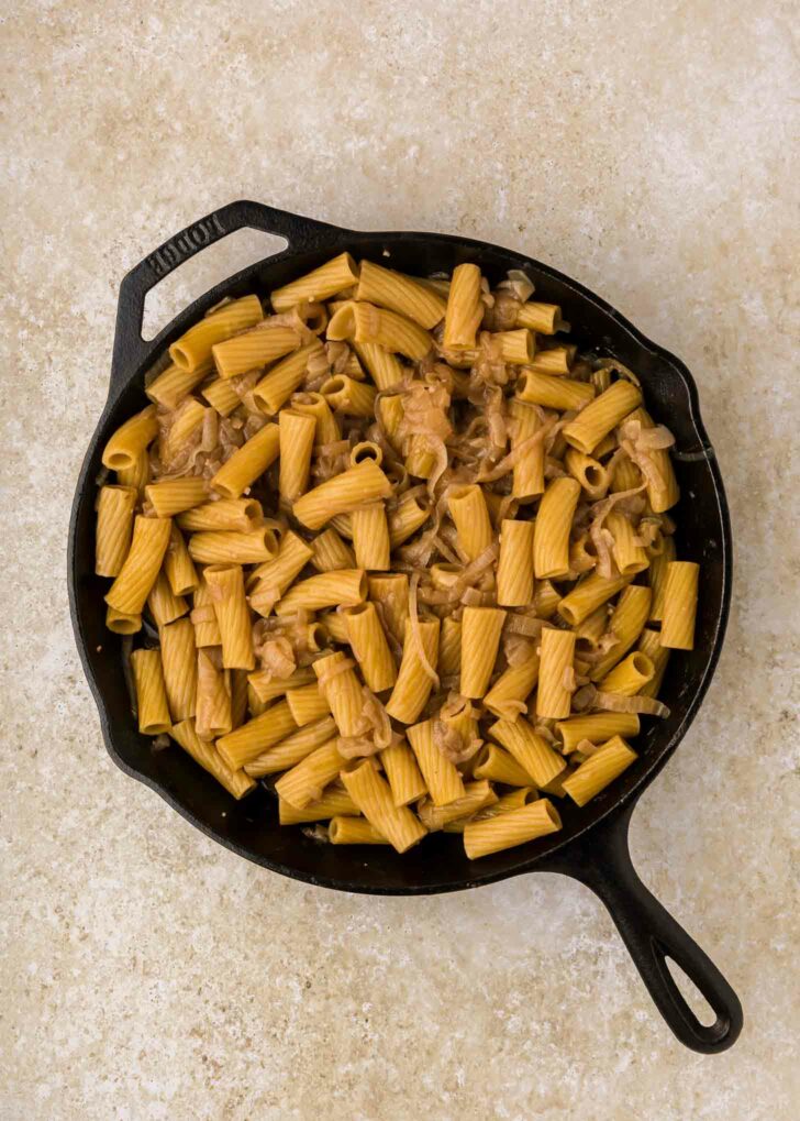 A black cast iron skillet filled with cooked rigatoni pasta mixed with caramelized onions, set on a light beige textured surface.