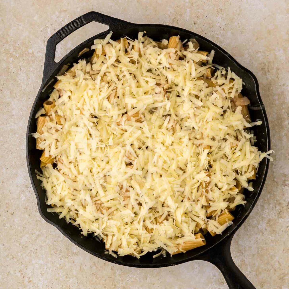 A black cast iron skillet filled with shredded cheese covering a layer of cooked ingredients, set on a light-colored countertop.