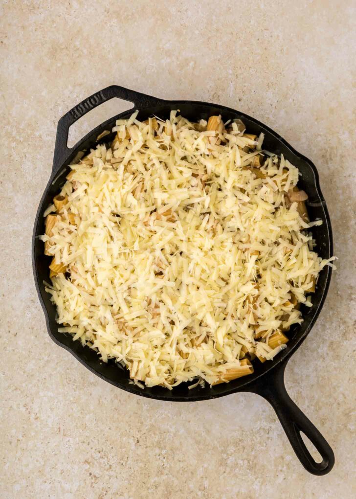 A black cast iron skillet filled with shredded cheese covering a layer of cooked ingredients, set on a light-colored countertop.
