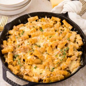 A cast iron skillet filled with rigatoni pasta with caramelized onions topped with melted cheese and fresh herbs, sitting on a light countertop next to plates, a block of cheese, and a white napkin.