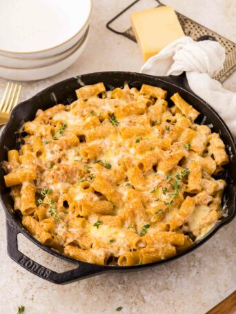 A cast iron skillet filled with rigatoni pasta with caramelized onions topped with melted cheese and fresh herbs, sitting on a light countertop next to plates, a block of cheese, and a white napkin.